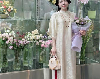 May include: A woman wearing a white dress with a red trim and a red beaded necklace. She is holding a bouquet of flowers and a white handbag with a gold chain.