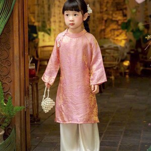 May include: A pink long-sleeved tunic with a subtle gold pattern, featuring a high collar and side button closure, paired with loose-fitting white trousers. The outfit is accessorized with a small white beaded handbag and a white hair accessory.