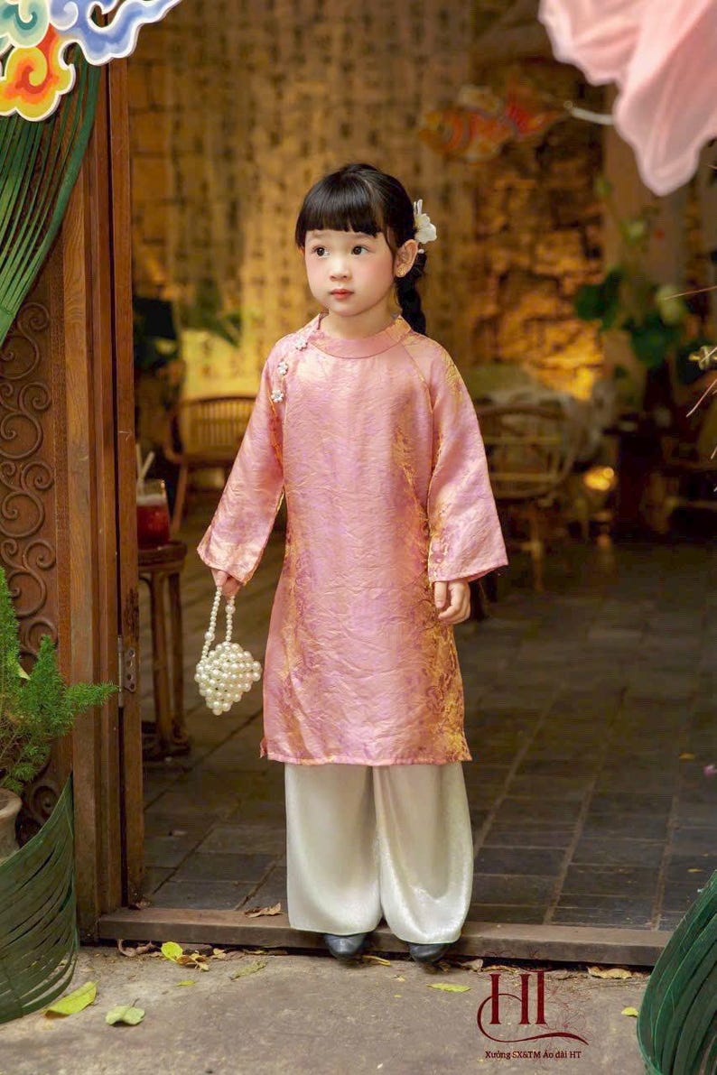 May include: A pink long-sleeved tunic with a subtle gold pattern, featuring a high collar and side button closure, paired with loose-fitting white trousers. The outfit is accessorized with a small white beaded handbag and a white hair accessory.