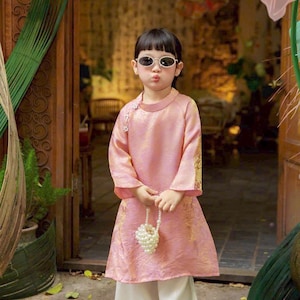 May include: A light pink long-sleeved tunic with a high collar and gold floral patterns, paired with loose white trousers. The tunic features a side closure with small white floral accents. A small handbag made of white faux pearls is held.