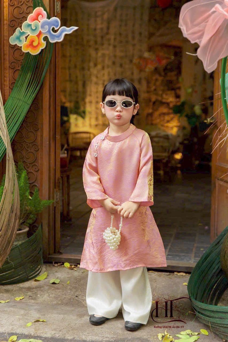 May include: A light pink long-sleeved tunic with a high collar and gold floral patterns, paired with loose white trousers. The tunic features a side closure with small white floral accents. A small handbag made of white faux pearls is held.