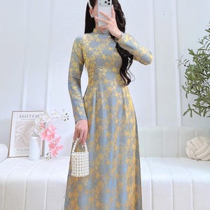 May include: A long, floor-length dress with long sleeves and a high collar. The dress has a floral pattern in yellow and grey. A small, white beaded purse is held. The background includes a white sofa and a framed print.