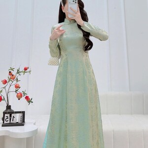 May include: A long, light green dress with a mandarin collar and long sleeves. The dress has a subtle floral pattern and a flowing silhouette. A small, white beaded purse hangs from the shoulder. The dress is suitable for formal occasions.