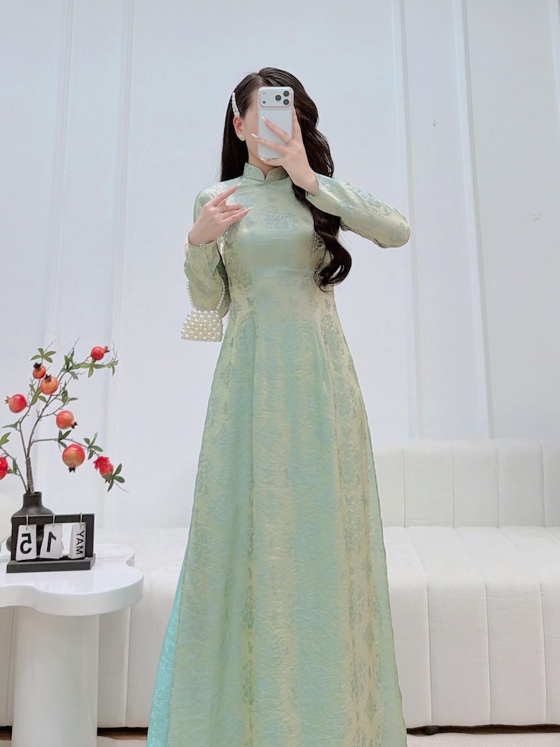 May include: A long, light green dress with a mandarin collar and long sleeves. The dress has a subtle floral pattern and a flowing silhouette. A small, white beaded purse hangs from the shoulder. The dress is suitable for formal occasions.