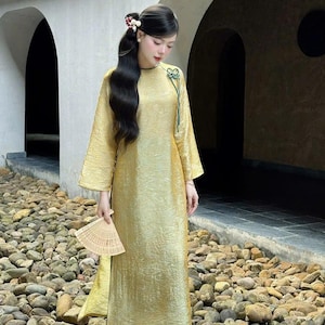 May include: A golden-yellow, long-sleeved dress with a textured appearance. The dress features a high neckline and a decorative green accent. The person is holding a beige fan. The dress is paired with teal-colored pants and white shoes.