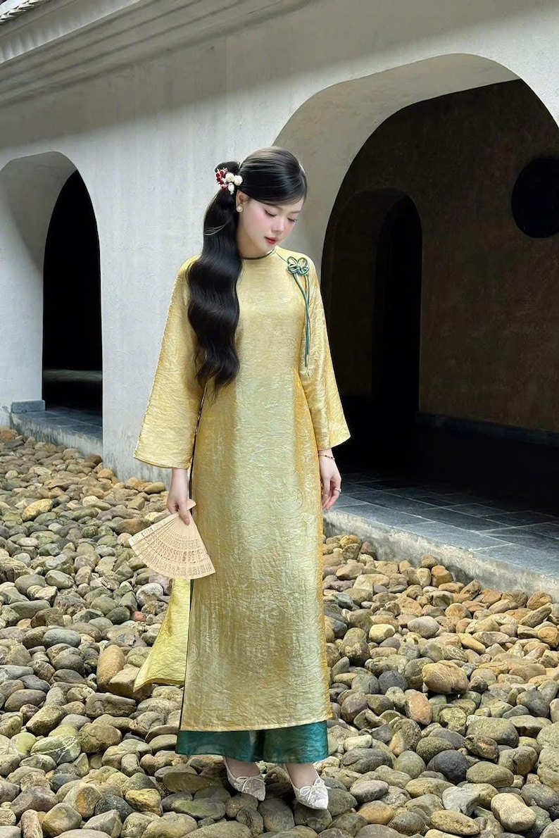 May include: A golden-yellow, long-sleeved dress with a textured appearance. The dress features a high neckline and a decorative green accent. The person is holding a beige fan. The dress is paired with teal-colored pants and white shoes.