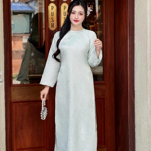 May include: A light green, long-sleeved dress with a floral pattern. The dress has a round neckline and a slit on the side. The person is holding a small, beaded purse. The background includes a wooden door with the word "PUSH" on it.