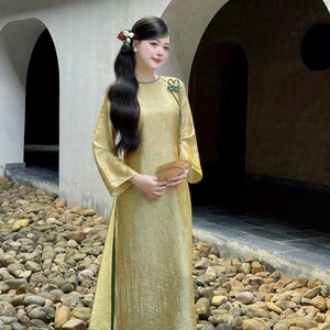 May include: A long, flowing gold dress with wide sleeves and a high slit. The dress has a green trim and is paired with green pants. A small fan is held in the hand. The background features stone and archways.