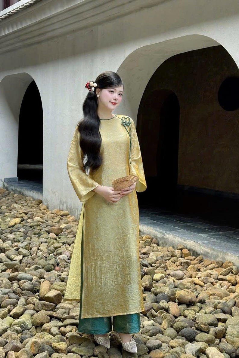 May include: A long, flowing gold dress with wide sleeves and a high slit. The dress has a green trim and is paired with green pants. A small fan is held in the hand. The background features stone and archways.