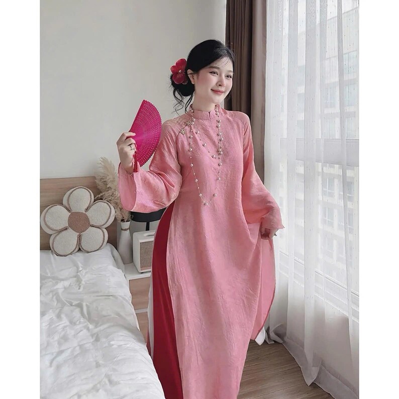 May include: A pink traditional dress with long sleeves and a high collar. The dress has a split on the side, revealing a red underlayer. A pink fan is held in the hand. The dress is adorned with delicate embroidery and a pearl necklace.