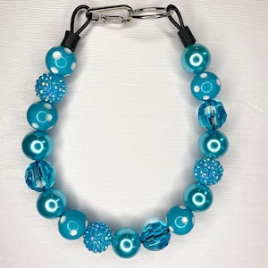 May include: A turquoise beaded necklace with a silver clasp. The necklace features a variety of beads, including solid, polka-dotted, and crystal-cut beads. The beads are strung together with a black cord.