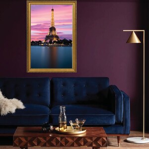 Puede incluir: Una fotografía enmarcada de la Torre Eiffel al atardecer, con un reflejo de la torre en el agua debajo. La foto está colgada en una pared morada sobre un sofá de terciopelo azul y una mesa de café de madera.