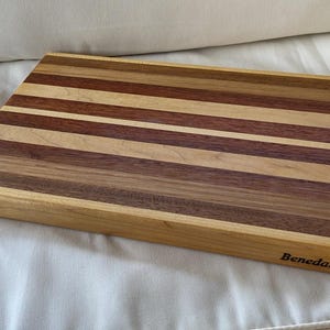 May include: A rectangular wooden chopping board featuring alternating horizontal stripes of light, medium, and dark brown wood tones. The board has a smooth, finished surface and the brand name Benedanti is clearly printed in black on the bottom right corner.
