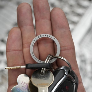 Puede incluir: Un llavero plateado con el texto "TI CORNER" grabado en él. El llavero está unido a un llavero de coche negro con tres botones y una llave plateada.