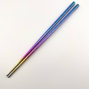 May include: A pair of rainbow-colored metal chopsticks. The chopsticks are made of stainless steel and have a gradient finish that goes from gold to purple.