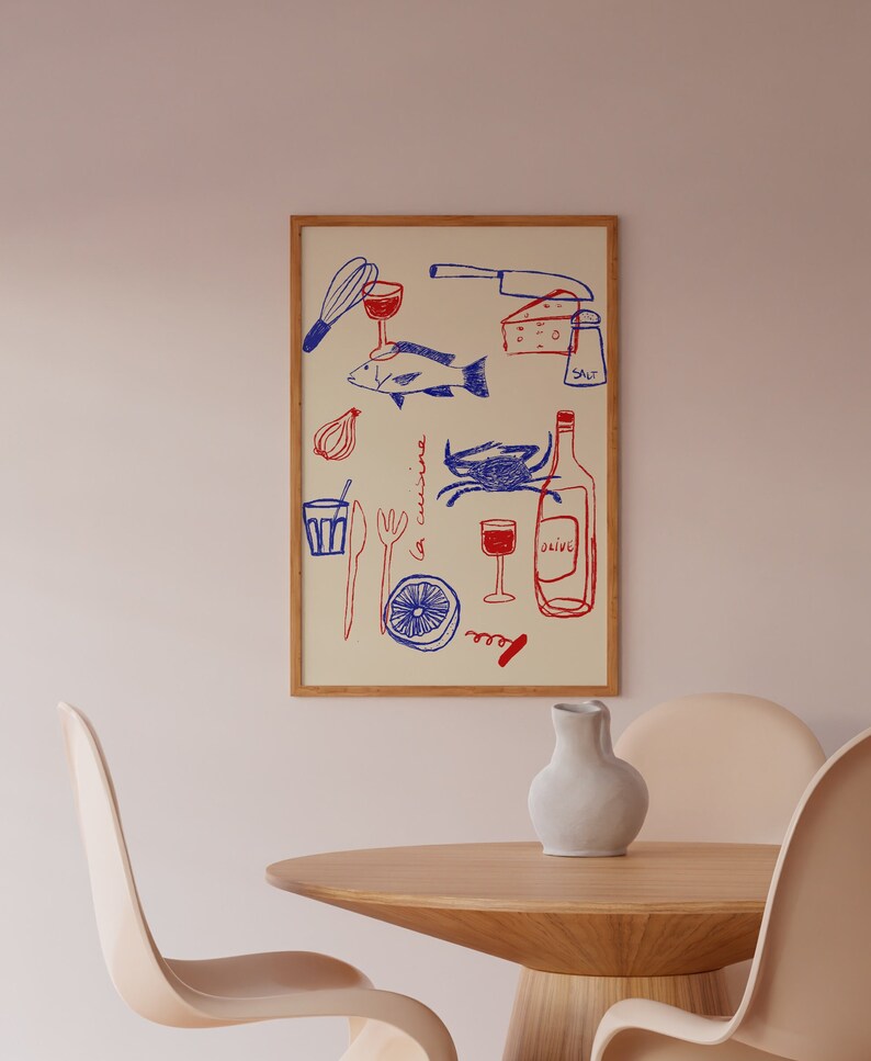 May include: A framed print with a white background and blue and red illustrations of various kitchen items, including a fish, a crab, a bottle of wine, a glass of wine, a knife, a whisk, a lemon, a fork, a spoon, a glass, a salt shaker, and the text "la cuisine" and "olive". The print is hanging on a light pink wall above a wooden table with two light brown chairs.