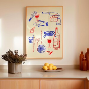 May include: A framed illustration of a kitchen scene with a red and blue color scheme. The illustration includes a fish, a crab, a bottle of wine, a glass of wine, a lemon, a whisk, a knife, a fork, a corkscrew, a bowl of onions, and a piece of cheese. The text "la cuisine" is written in blue on the illustration.