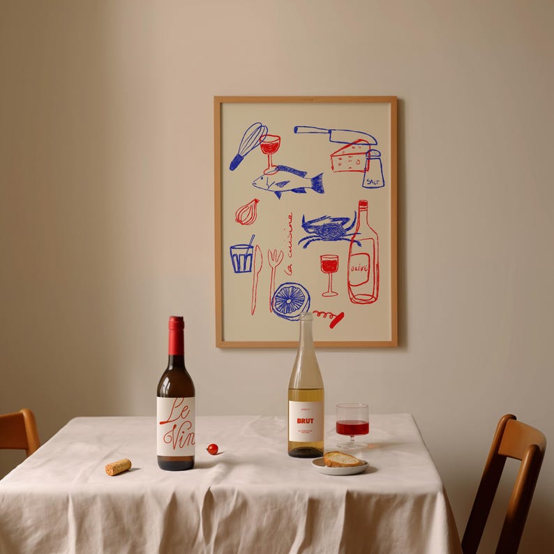 May include: A white tablecloth with two bottles of wine, one red and one white, and a glass of red wine. There is a framed print on the wall behind the table with a blue and red illustration of kitchen items, including a fish, a crab, a knife, and a glass of wine. The print has the text "la cuisine" in red.