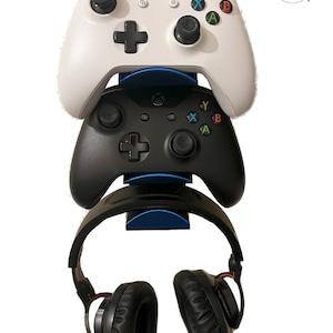 May include: A blue wall mount with two black and white Xbox controllers and a black headset hanging from it. The controllers are stacked on top of each other.