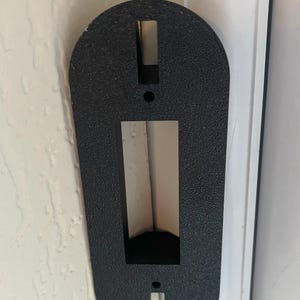 May include: Black plastic doorstop with a rectangular opening and two small holes. The doorstop is mounted on a white door frame.