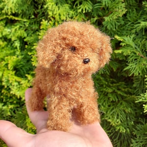 Op de afbeelding: Een kleine, bruine, gehaakte speelgoedpoedel met zwarte ogen. Het speelgoed zit op de hand van een persoon.