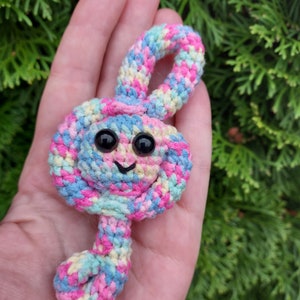 May include: A colorful crocheted treble clef with a smiling face and black eyes. The clef is made with pink, blue, yellow, and green yarn.