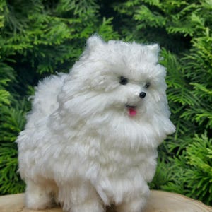 Puede incluir: Figura de perro Pomerania blanco con lengua rosa, colocada sobre una rodaja de madera sobre un fondo de follaje verde. El perro de juguete tiene ojos negros y nariz negra. El pelaje del perro es esponjoso.