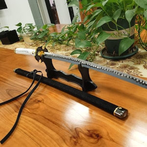 May include: A katana sword with a white handle, a gold-colored guard, and a grey blade featuring a serrated edge. The sword rests on a black stand next to a black scabbard with a long black cord attached to the end, showcasing a detailed design.