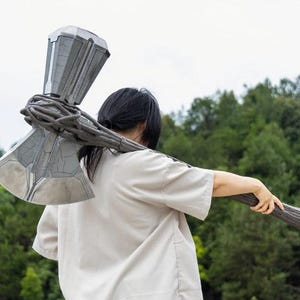 May include: A large battle axe with a metallic silver head featuring intricate panel lines and a dark, textured handle that resembles twisted wood or vines, held over the shoulder. The axe head has a wide, flat blade on one side and a blocky, rectangular hammer-like structure on the top, connected by a series of dark, vine-like wrappings.