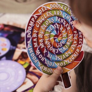 May include: A colorful, circular game piece shaped like a lollipop with the words "DOOMPA DEE DOO! I'VE GOT MANY GIGGLES ARE THERE YOU! HOW MARVELOUS FINGER SURPRISE INKING CHO- SOLA ADVISE FOR A RIDDLE PLEASE PERFECT" printed on it.