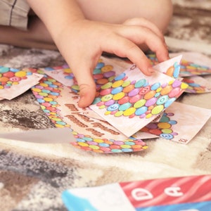 May include: A child's hand holding a colorful card game with a candy theme. The card features a variety of colorful candies and the text "The Candy Fruit & Nut Game".
