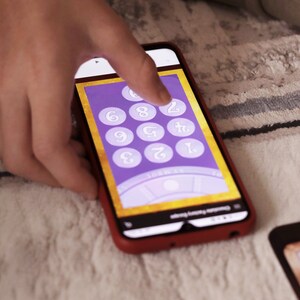 May include: A smartphone displaying a purple and gold digital game board with numbers 1 through 9. The game board is on a mobile phone screen with a red border.