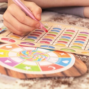 May include: A colorful game board with a grid of circles in various colors. A child's hand is holding a pink marker and is marking a circle on the board. The game board is made of cardboard and has a wooden frame. There is a smaller circular game piece with a colorful spiral design.