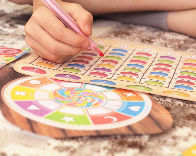 May include: A colorful game board with a grid of circles in various colors. A child's hand is holding a pink marker and is marking a circle on the board. The game board is made of cardboard and has a wooden frame. There is a smaller circular game piece with a colorful spiral design.