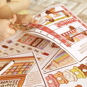May include: A child's hands using scissors to cut out a colorful activity sheet. The sheet features illustrations of ice cream cones, donuts, and other treats. Other sheets with word puzzles and a marker are also visible.