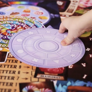 May include: A purple and white circular game piece with a star, crescent moon, and other symbols. The piece is on a game board with a colorful background and the words "For You", "Ise In", "Choc", "Sur", "Angel", "Giggles", "Doth", and "Ego" visible.