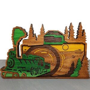 May include: A wooden cutout of a camera with a train traveling through the lens. The camera is painted in shades of brown, yellow, and green. The train is green with black accents. The background is a forest scene with green trees.