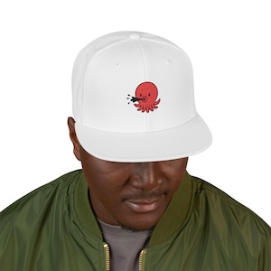 May include: A white snapback cap featuring an embroidered graphic of a small, round, red octopus with an angry expression, shown spitting a stream of black ink from its mouth. The cap has a structured crown and a flat brim, centred on the front.