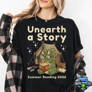 May include: Black t-shirt with the text "Unearth a Story" above a cartoon dinosaur reading a book in front of a tent. The phrase "Summer Reading 2026" is below the dinosaur illustration. The shirt has a digital download graphic.