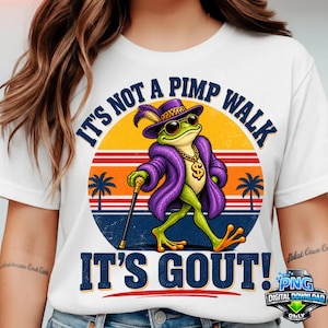 Op de afbeelding: Een wit t-shirt met een afbeelding van een groene kikker in een paarse bontjas, een paarse hoed met veer, een zonnebril, een gouden ketting met dollarteken en een wandelstok. De tekst luidt IT'S NOT A PIMP WALK IT'S GOUT! boven een zonsondergang.
