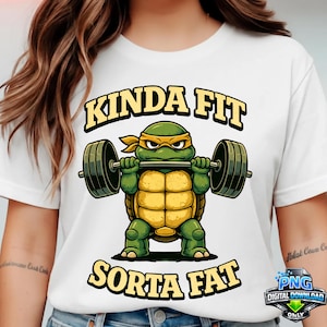 Op de afbeelding: Wit t-shirt met een cartoon schildpad die gewichten heft. De schildpad is groen en geel en draagt een gele hoofdband. De tekst op het shirt luidt "KINDA FIT" boven de schildpad en "SORTA FAT" eronder.