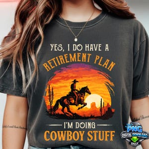 May include: Dark gray t-shirt with a sunset graphic featuring a cowboy on a horse. Text reads "YES, I DO HAVE A RETIREMENT PLAN - I'M DOING COWBOY STUFF."