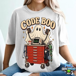 Op de afbeelding: Een wit t-shirt met een afbeelding van een geest die twee luidsprekers boven een rode medische kar houdt. De kar bevat een hartmonitor met een groene pulslijn, een stethoscoop, een groene zuurstoffles en een bloeddrukmeter. De tekst CODE BOO staat erboven.