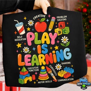 May include: Black t-shirt with a colorful graphic that reads "PLAY IS LEARNING." The design includes illustrations of an apple, pencil, building blocks, and other educational elements. The shirt also features the words "Creative," "Problem Solving," and "Motor Skills."