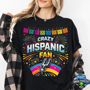 Puede incluir: Camiseta negra con el texto "CRAZY HISPANIC FAN" en un diseño colorido y festivo. La camiseta presenta fuegos artificiales, un arcoíris y una pancarta de papel picado. Un logotipo de descarga digital está en la esquina inferior derecha.