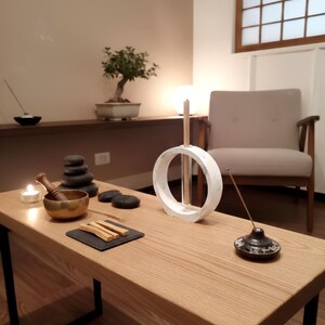 May include: A wooden table with black metal legs displays spa and meditation items. A white circular sculpture, incense, a brass bowl, and a stack of black stones are arranged on the table. A bonsai tree sits on a shelf in the background.