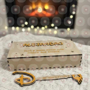 Puede incluir: Una caja de madera con el nombre "ALEJANDRO" en letras naranjas. La caja tiene texto en español y está acompañada de una llave decorativa con el texto "NOS VAMOS A DISNEY !!". La llave y la caja están sobre una superficie blanca.