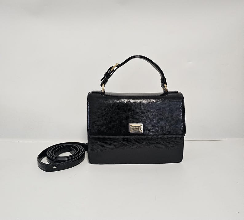 K&ouml;nnte beinhalten: Eine schwarze Handtasche mit strukturierter Oberfl&auml;che, mit einem oberen Griff und einem abnehmbaren Schulterriemen. Die Tasche hat eine rechteckige Form mit einem Klappverschluss und einem kleinen silberfarbenen Metalldetail auf der Vorderseite. Der Riemen ist schwarz.