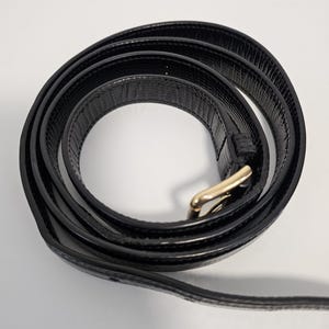 K&ouml;nnte beinhalten: Ein aufgerollter schwarzer Lederg&uuml;rtel mit einer goldfarbenen Schnalle. Der G&uuml;rtel hat eine strukturierte Oberfl&auml;che und wird vor einem wei&szlig;en Hintergrund gezeigt. Die Schnalle ist rechteckig mit abgerundeten Kanten.