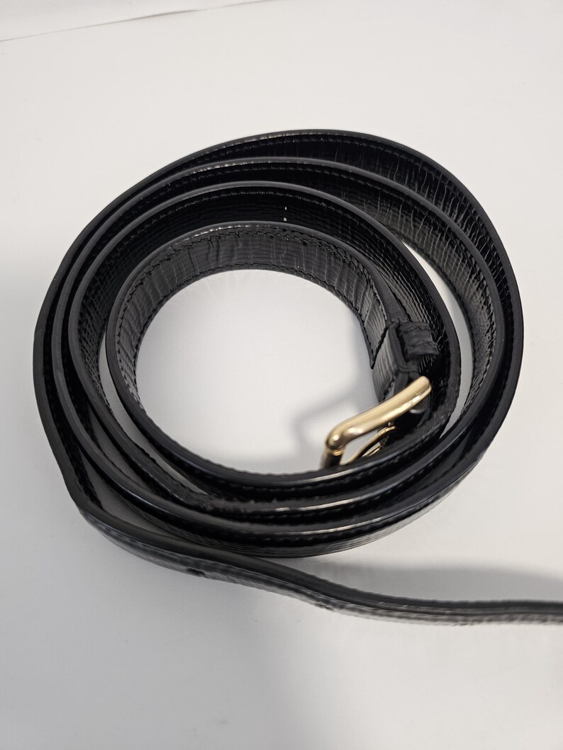 K&ouml;nnte beinhalten: Ein aufgerollter schwarzer Lederg&uuml;rtel mit einer goldfarbenen Schnalle. Der G&uuml;rtel hat eine strukturierte Oberfl&auml;che und wird vor einem wei&szlig;en Hintergrund gezeigt. Die Schnalle ist rechteckig mit abgerundeten Kanten.